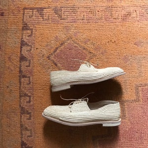 K&ouml;nnte beinhalten: Zwei Paar wei&szlig;e gewebte Schuhe mit Schn&uuml;rsenkeln auf einem braunen gemusterten Teppich.