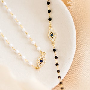 May include: Two gold necklaces with evil eye pendants. One necklace has white beads and the other has black beads.