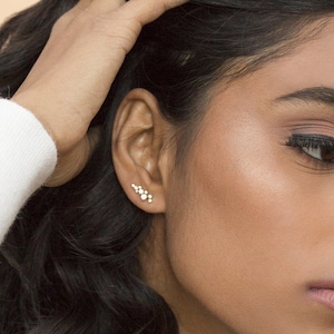 May include: A woman with long, dark, curly hair wearing a white sweater and a gold necklace with a small star charm. She is wearing a pair of small, gold earrings with a cluster of diamonds.