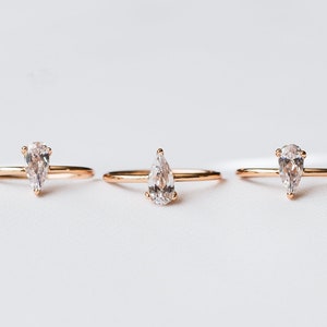 Może przedstawiać: Trzy złote pierścionki z przezroczystymi kamieniami szlachetnymi w kształcie gruszki. Pierścionki są ułożone w rzędzie na białym tle.