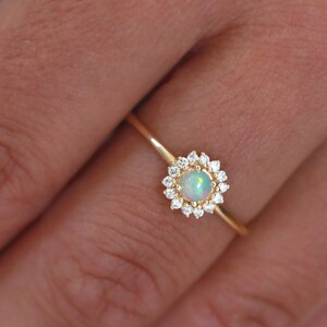 Könnte beinhalten: Ein zarter Goldring mit einem runden Opal-Edelstein, der von einem Heiligenschein aus kleineren Diamanten umgeben ist.