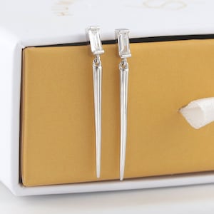 Könnte beinhalten: Ein Paar silberne Ohrringe mit einem langen, dünnen, spitzen Design. Jeder Ohrring hat oben einen kleinen, rechteckigen, klaren Stein.