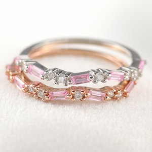 May include: Two delicate rings with a wavy design, featuring alternating pink and clear gemstones. The rings are displayed on a white velvet box.