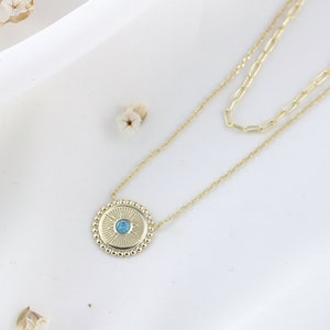 May include: A gold necklace with a round pendant featuring a blue gemstone surrounded by a beaded border. The pendant has a sunburst design.