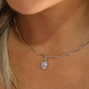 Op de afbeelding: Een zilverkleurige ketting met een fijne, golvende schakelketting en een enkele ovaalvormige hanger met een heldere, gefacetteerde steen in een gladde metalen zetting. De hanger hangt centraal aan de getextureerde, golvende ketting.