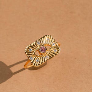May include: A gold-colored ring with an eye-shaped design. The ring features a pink center stone and small white stones around the eye. The ring is set against a neutral background with a leaf shadow.