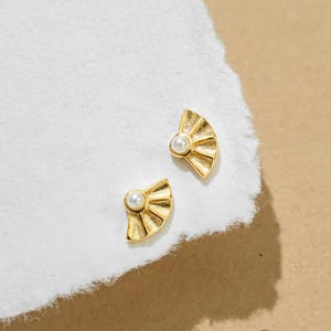 May include: Two gold-colored fan-shaped stud earrings with a small, round, white pearl in the center of each. The earrings are displayed on a torn piece of white paper, set against a tan background.