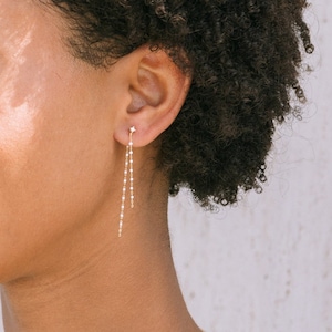 Puede incluir: Una mujer con el pelo corto y rizado castaño lleva una blusa blanca con un collar de oro y pendientes a juego de oro. Los pendientes son largos y cuelgan con una cadena de pequeñas cuentas.