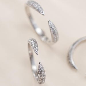Op de afbeelding: Drie open zilveren ringen, elk bezet met kleine, sprankelende, heldere stenen. De ringen zijn gerangschikt op een lichtbeige oppervlak, wat een minimalistische en elegante esthetiek creëert. Het open ontwerp voegt een moderne touch toe aan de sieraden.