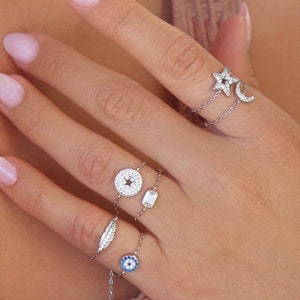 Puede incluir: Una mano con varios anillos delicados de cadena plateada con pequeños colgantes, incluyendo una estrella, una luna, un ojo y un dije redondo con pequeñas piedras. Los anillos están en un dedo con esmalte de uñas rosa claro.