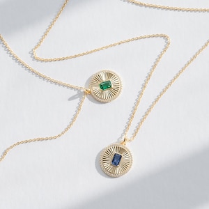 May include: Two gold necklaces with pendants. One pendant features a green gemstone in a sunburst design. The other pendant features a blue gemstone in a sunburst design.