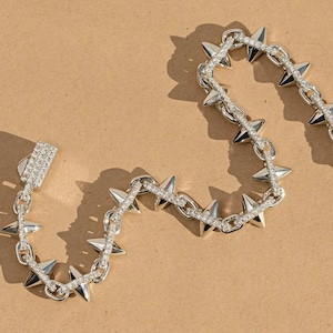 May include: A silver bracelet with a spiky design. The bracelet has alternating spikes and small, clear stones. The clasp is also decorated with small, clear stones. The bracelet is shown on a tan surface.