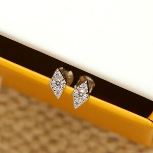 May include: A pair of diamond-shaped stud earrings with small, sparkling stones. The earrings are displayed inside a white box with a yellow interior and a white ribbon pull. The box has gold lettering on the top.