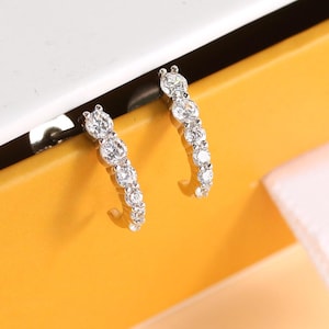 May include: A pair of silver hoop earrings with clear, sparkling stones. The earrings are displayed on a yellow and white box with the word "HUM" visible. A white ribbon is attached to the box.