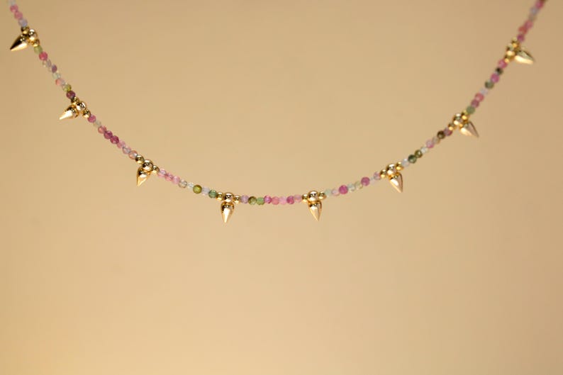 May include: A delicate necklace featuring a strand of small, colorful beads in shades of pink, green, and white. Gold-colored, pointed accents are evenly spaced along the necklace, creating a unique and elegant design.