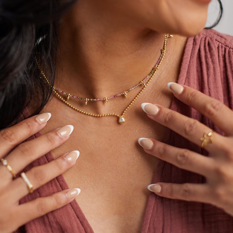 May include: A layered gold necklace with a small pendant and beaded chain. The top layer features small pink beads and gold accents. The image also shows gold rings on the fingers and a mauve-colored top.