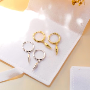 May include: Two pairs of hoop earrings, one pair in gold tone and one pair in silver tone. Each hoop features a small, pointed, cone-shaped charm dangling from the bottom. The earrings are arranged on a white fabric surface with a soft texture.