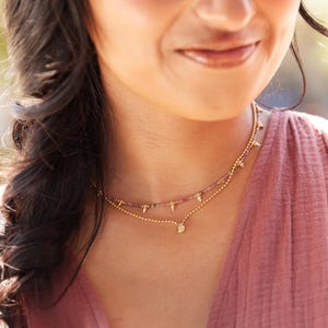 May include: A layered gold necklace with a delicate design. The top layer features small pink and purple beads, while the lower layer has a small pendant. The necklace has a simple, elegant style.