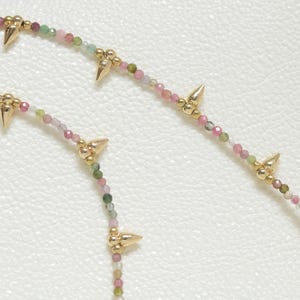 May include: Close-up of two necklaces with small, colorful beads in shades of pink, green, and white. The necklaces feature gold-colored spike accents. The background is a textured, off-white surface.