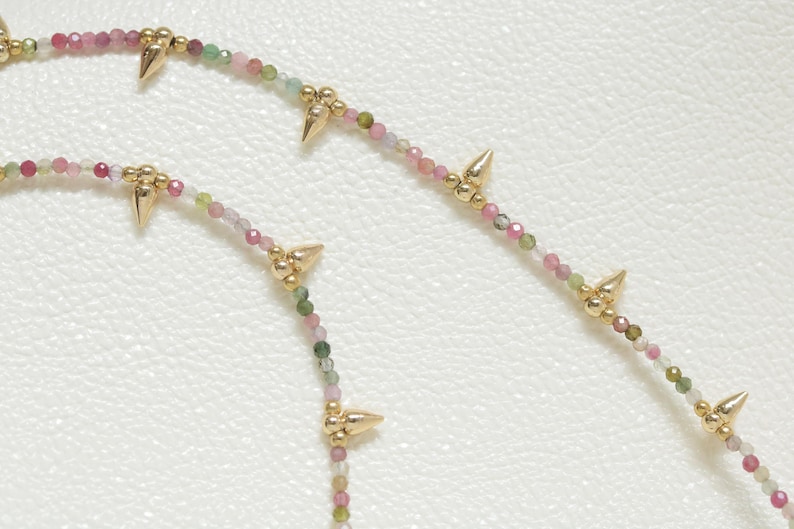 May include: Close-up of two necklaces with small, colorful beads in shades of pink, green, and white. The necklaces feature gold-colored spike accents. The background is a textured, off-white surface.