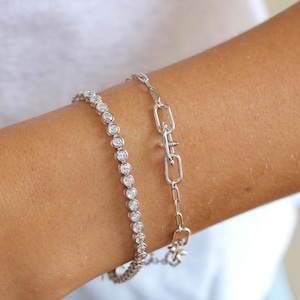 May include: A silver-toned bracelet featuring a continuous row of round, bezel-set clear stones alongside a second silver-toned bracelet composed of elongated rectangular chain links with a central toggle-style clasp mechanism.