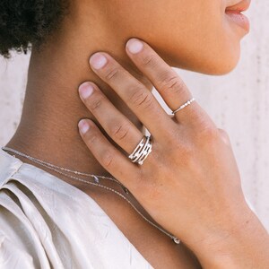 May include: A woman wearing a white top with a delicate silver necklace and a silver ring with multiple bands. The ring is adorned with small, sparkling stones.