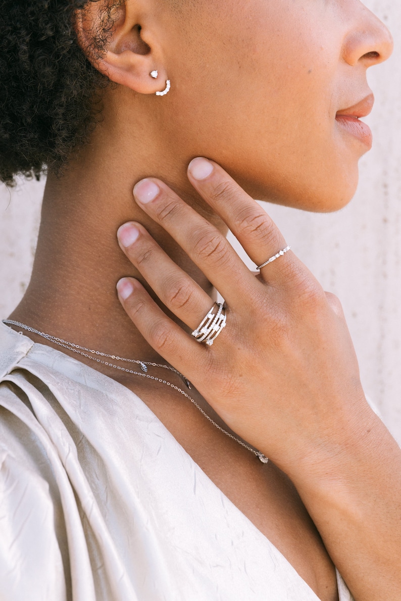 May include: A woman wearing a white top with a delicate silver necklace and a silver ring with multiple bands. The ring is adorned with small, sparkling stones.