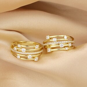 May include: Two gold rings with multiple bands and small clear stones. The rings are on a beige fabric background.
