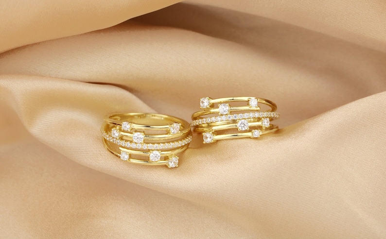 May include: Two gold rings with multiple bands and small clear stones. The rings are on a beige fabric background.
