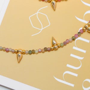May include: A delicate necklace featuring small, multi-colored beads in shades of pink, green, and gold. The necklace is accented with gold-colored, cone-shaped pendants. The background is a light yellow with white text.