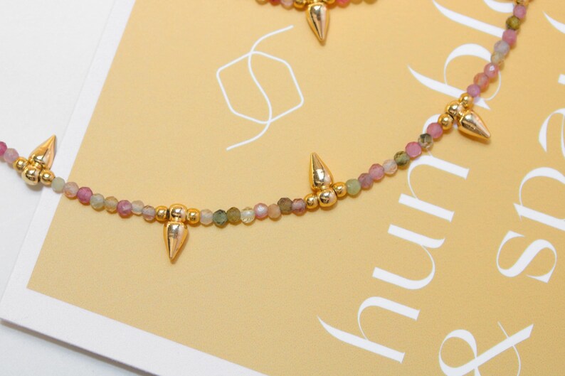 May include: A delicate necklace featuring small, multi-colored beads in shades of pink, green, and gold. The necklace is accented with gold-colored, cone-shaped pendants. The background is a light yellow with white text.