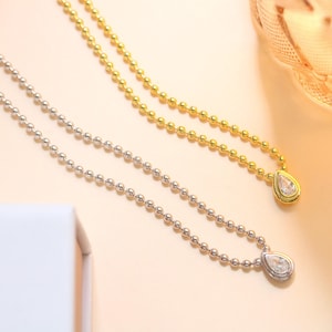 May include: Two delicate necklaces with teardrop pendants. One necklace is gold-toned, the other is silver-toned. Both feature a chain of small, round beads. The pendants have a clear, sparkling stone set in a bezel.