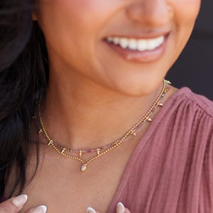 May include: A gold necklace with two strands. The top strand has small gold spikes and the bottom strand has a teardrop pendant. The necklace also has small colorful beads. The person in the image is wearing a pink top.