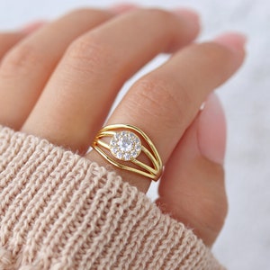 Könnte beinhalten: Ein goldener Ring mit einem einzigen runden, klaren Stein, der von kleineren Steinen umgeben ist. Der Ring hat drei Bänder, die leicht versetzt zueinander sind.