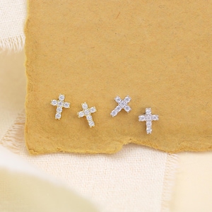 May include: Four small, silver-colored cross-shaped earrings with clear gemstones are arranged on a textured, tan-colored paper. The earrings are displayed on a neutral background with a soft, off-white fabric in the lower left corner.