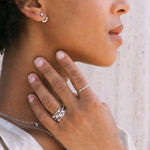 May include: A woman's hand wearing a silver ring with multiple small diamonds and a silver ring with a single row of small diamonds. The woman is wearing a silver necklace and a silver earring with a small diamond.