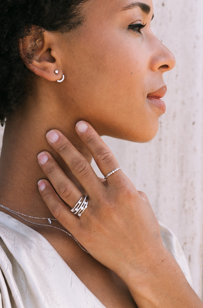 May include: A woman's hand wearing a silver ring with multiple small diamonds and a silver ring with a single row of small diamonds. The woman is wearing a silver necklace and a silver earring with a small diamond.