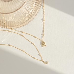May include: Two gold-colored necklaces with delicate chains and small beaded accents. One necklace features the letter "R" as a pendant, and the other has the letter "L". The necklaces are displayed on a white surface.