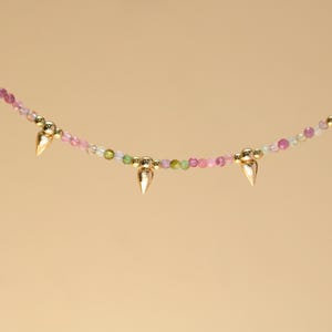 May include: A delicate necklace featuring a strand of small, colorful beads in shades of pink, green, and clear. Gold-colored cone-shaped pendants are evenly spaced along the necklace. The background is a solid, light tan color.
