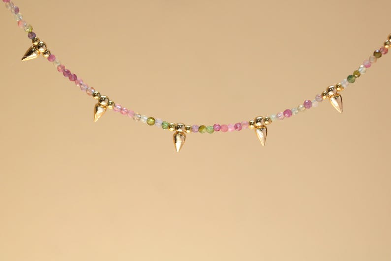May include: A delicate necklace featuring a strand of small, colorful beads in shades of pink, green, and clear. Gold-colored cone-shaped pendants are evenly spaced along the necklace. The background is a solid, light tan color.