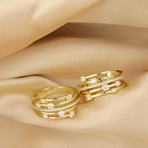 May include: Two gold rings with multiple bands and clear cubic zirconia stones. The rings are on a beige fabric background.