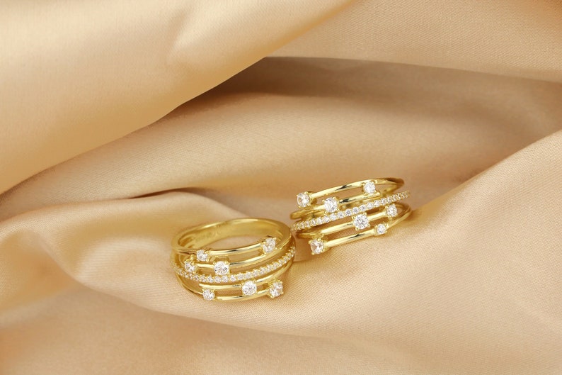 May include: Two gold rings with multiple bands and clear cubic zirconia stones. The rings are on a beige fabric background.