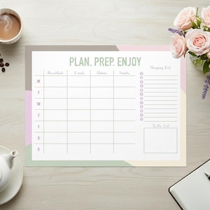 May include: A weekly meal planning notepad with the words "PLAN. PREP. ENJOY" at the top. The notepad has sections for each day of the week, a shopping list, and a to-do list. The notepad is on a light wood surface.