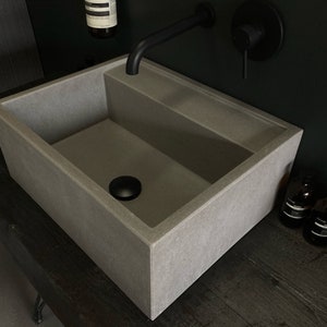 May include: A rectangular concrete sink with a black drain stopper. The sink is mounted on a dark wood countertop. A black faucet is mounted on the wall behind the sink.