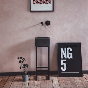 May include: A black framed picture with a geometric pattern of red, white and black triangles hangs on a pink wall above a black sink on a black stand. A small potted plant sits on the wooden floor. A black framed picture with the text "NG 5" is on the right side of the image.