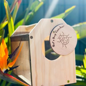 May include: A wooden bug catcher with a round, engraved lid featuring a ladybug design and the text "Sal's Bug Catcher." The catcher has a mesh side and a circular opening. The wood is light-colored, and the screws are silver and green.