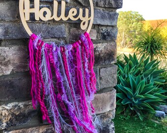 May include: A wooden hoop with the name "Holley" written on it. The hoop is decorated with pink and purple yarn and fabric.