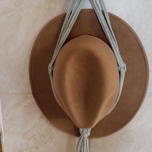 May include: A brown hat suspended by a macramé hat hanger. The hanger is made of light blue rope and a wooden dowel, adorned with dried flowers and an orange flower. The hat has a wide brim.