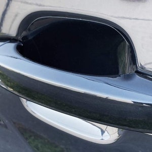 Puede incluir: Una manija de puerta de coche negra con un acento cromado. La manija está empotrada en la puerta y tiene un pequeño botón en el lateral.