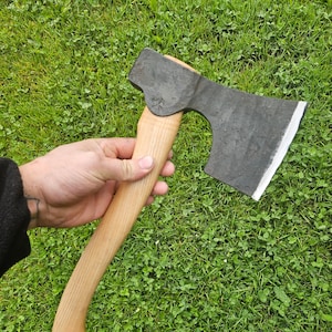 May include: A hand holds a black axe with a wooden handle. The axe has a curved blade and a slightly rounded head. The axe is set against a background of green grass.